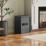 A black electronic home safe standing on a polished wood floor next to a fireplace and a potted plant.